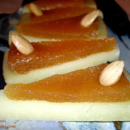 This horizontal rectangle photo shows the completed Membrillo (Quince Paste) recipe by Reese Amorosi for GlamorosiCooks.com. There is a row of triangle-shaped appetizers made of Manchego cheese topped with the membrillo (quince paste) then garnished with Marcona almonds. The appetizers are arranged in a row on a blue abstract pottery plate that is in a vertical position through the center of the photo. The sides of the plated are a muted pink - it is a small 50's Formica dinette table.