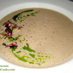 This photo shows the completed Roasted Cauliflower Soup recipe by Reese Amorosi of GlamorosiCooks.com. The soup is in a white bowl with wide, flare sides. The soup is light tan. It is garnished with roasted cauliflower florets, green pumpkin seeds, red pomegranate seeds and green basil oil. The garnish is in a right-facing crescent moon shape on the left side of the bowl. Recipe and photo copyright Reese Amorosi GlamorosiCooks.com