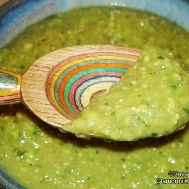 The photo shows the completed Salsa Verde with Avocado recipe by Reese Amorosi for GlamorosiCooks.com. It is a close-up shot of a blue ceramic bowl of Salsa Verde. A wooden spoon with a rainbow pattern contains salsa verde and is held above the bowl.