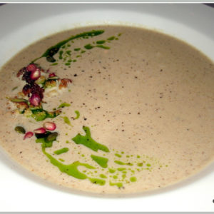 This photo shows the completed Roasted Cauliflower Soup recipe by Reese Amorosi of GlamorosiCooks.com. The soup is in a white bowl with wide, flare sides. The soup is light tan. It is garnished with roasted cauliflower florets, green pumpkin seeds, red pomegranate seeds and green basil oil. The garnish is in a right-facing crescent moon shape on the left side of the bowl. Recipe and photo copyright Reese Amorosi GlamorosiCooks.com