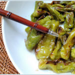 This photo shows the completed Roasted Shishito Peppers with Honey and Smoked Sea Salt recipe. The peppers are green and slightly charred, and they are glossy from the honey glaze. They are in a white bowl on a rattan charger, and there is a brown bamboo fork reaching into the food. Photo and Recipe copyright Reese Amorosi of Glamorosi Cooks