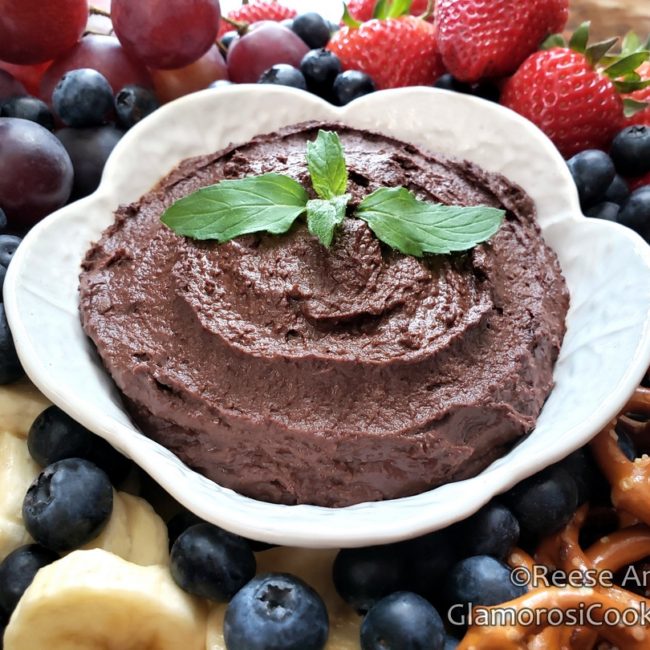 This photo is a horizontal rectangle, and shows the completed Chocolate Hummus recipe by Reese Amorosi for GlamorosiCooks.com. The Chocolate Hummus is in a white vintage ceramic bowl that looks like half of a hollowed out cabbage, and it is garnished with peppermint leaves. The bowl is surrounded by a variety of items to dip in the Chocolate Hummus: clockwise from the bottom left there are sliced bananas, red seedless grapes, strawberries and mini hard pretzels. There are blueberries encircling the entire bowl.