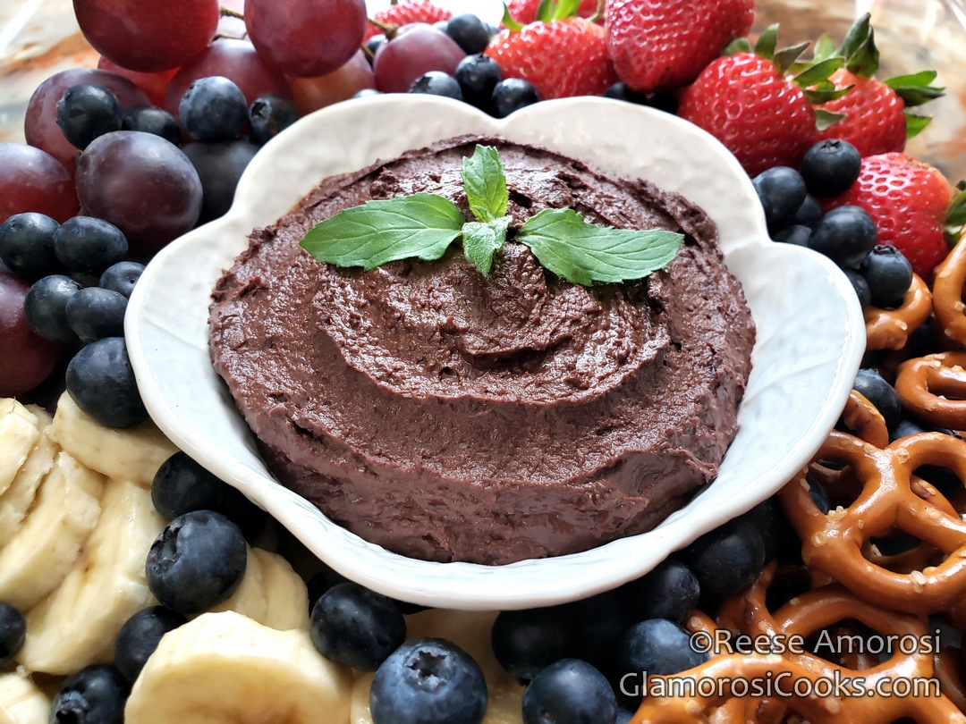 This photo is a horizontal rectangle, and shows the completed Chocolate Hummus recipe by Reese Amorosi for GlamorosiCooks.com. The Chocolate Hummus is in a white vintage ceramic bowl that looks like half of a hollowed out cabbage, and it is garnished with peppermint leaves. The bowl is surrounded by a variety of items to dip in the Chocolate Hummus: clockwise from the bottom left there are sliced bananas, red seedless grapes, strawberries and mini hard pretzels. There are blueberries encircling the entire bowl.