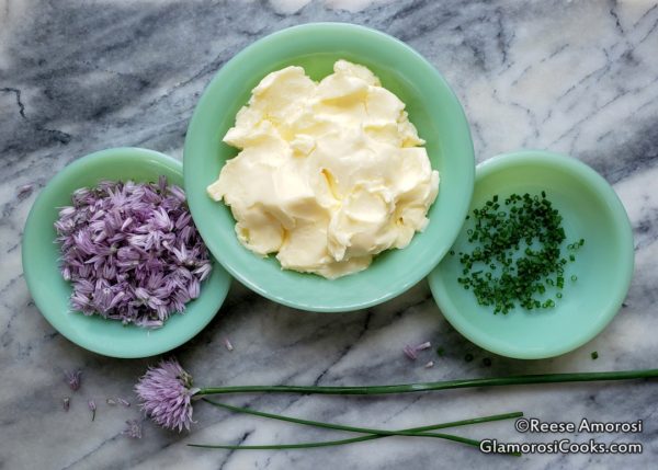This photo shows Chive Blossom Butter Ingredients - from left to right: chive flowers separated, unsalted butter, chopped chive leaves. The ingredients are in jadeite bowls, large in the middle, smaller on either side. There is a whole chive flower and two chive leaves in front of the bowls. All of it is on a grey marble tray hat fills the frame. Recipe and Photo Copyright Reese Amorosi, GlamorosiCooks.com