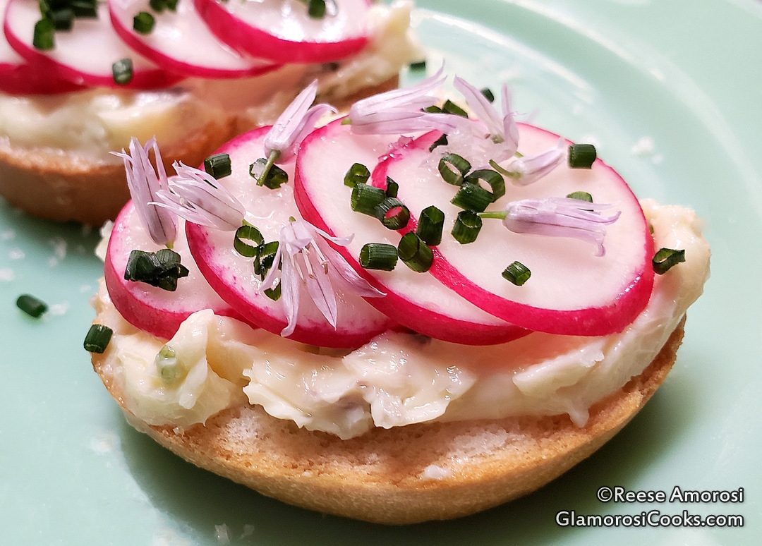 This is a photo of Chive Blossom Butter Radish Toast. The butter is on the toast and it is topped with sliced radishes that have pink skin and white flesh. The radishes are topped with chopped green chives and pretty lavender chive flowers. The radish toast is sitting on a Jadeite plate. Recipe and Photo Copyright Reese Amorosi, GlamorosiCooks.com