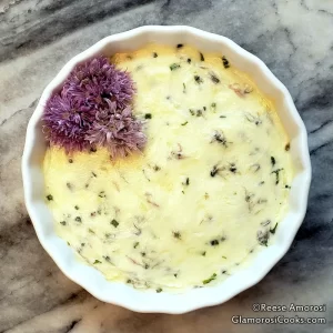 This is a photo of the finished Chive Blossom Butter recipe. The butter is yellow and flecked with bits of purple chive blooms and chopped green chive leaves. The butter is in a round white bowl with fluted edges and there are three whole chive blooms garnishing the butter - they are in the 11 o'clock position. All of it is on a grey marble tray that fills the whole frame. Recipe and Photo Copyright Reese Amorosi, GlamorosiCooks.com