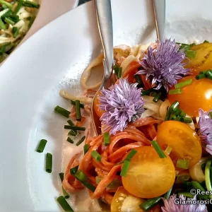 This is a photo of sundried tomato, squash and spinach fettuccini. It is garnished with orange Sungold tomatoes, chopped green chives and puffy purple chive blossoms. There is also alfredo sauce. Chive Blossom Pasta - Recipe and Photo Copyright Reese Amorosi, GlamorosiCooks.com