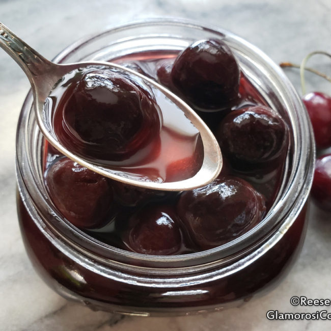This photo shows the completed recipe for Bourbon Soaked Cherries by Reese Amorosi for GlamorosiCooks.com. The square photo shows an overhead, close up view of the Bourbon Soaked cherries in a clear canning jar. A silver spoon containing one cherry hovers over the open jar, and a few whole cherries are next to the jar. The background is grey marble.