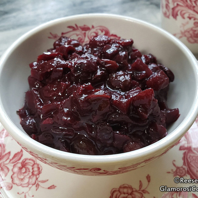 This photo shows the completed Cherry Chutney recipe by Reese Amorosi for GlamorosiCooks.com. The chutney is in a white bowl with red floral designs, and the bowl is on a matching plate next to a partially visible matching cup. All of it sits on a grey marble serving tray.