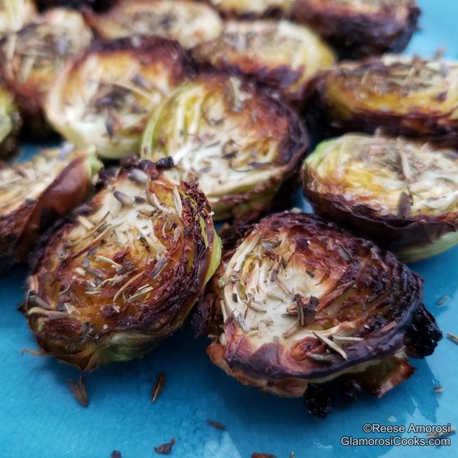 This photo shows a close-up of the completed Roasted Brussels Sprouts with Herbes de Provence recipe by Reese Amorosi for GlamorosiCooks.com. seasoned with The Brussels Sprouts are on a blue ceramic plate. They are cut in half and are crispy and caramelized from being roasted. The Herbes de Provence are visible on the vegetables, and some has fallen off onto the plate.