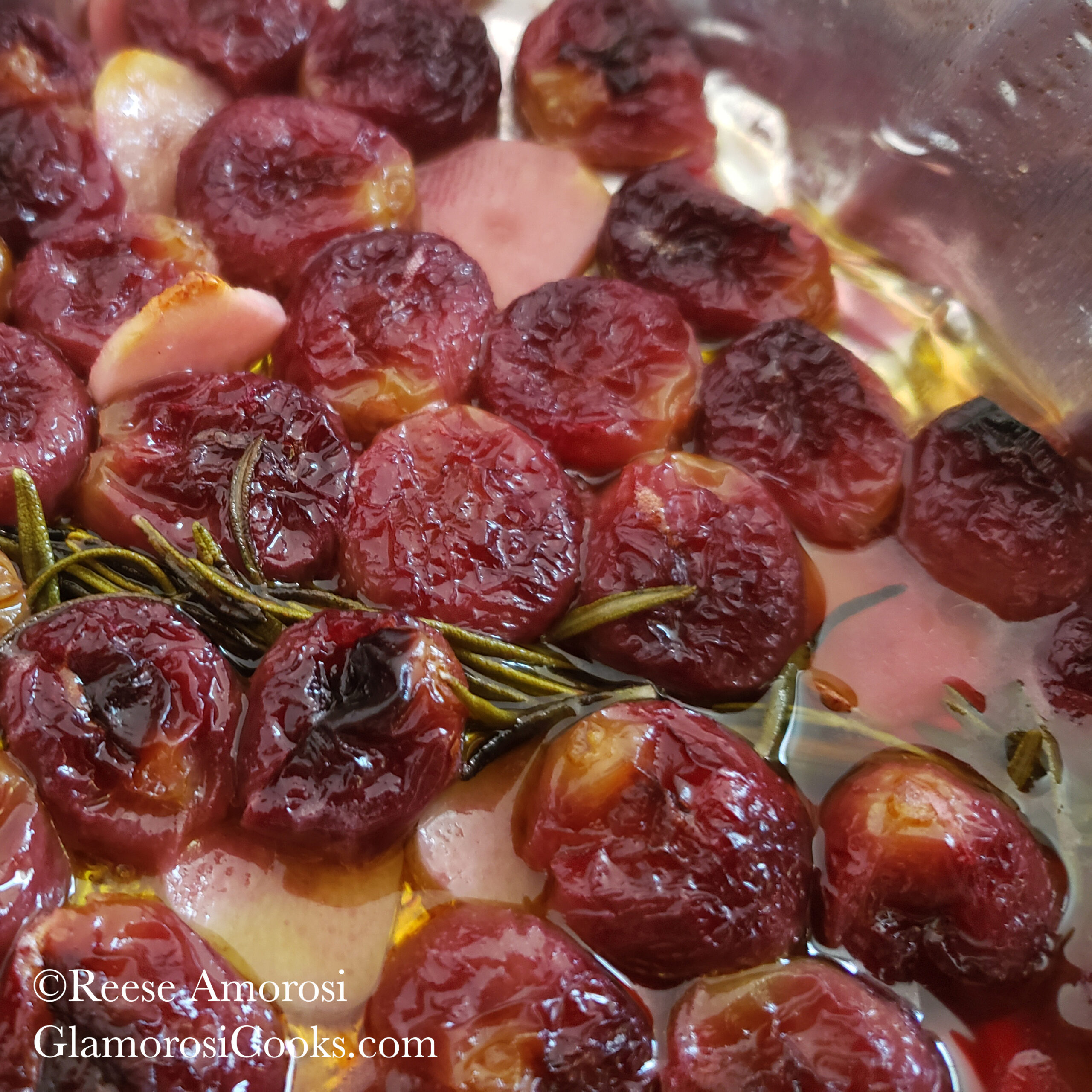 This photo shows the completed Roasted Grapes recipe by Reese Amorosi of GlamorosiCooks.com. The seedless red grapes are charred on purpose, as that is part of what makes the flavor so good. The grapes are in a shallow bowl that is barely visible due to how close the photo was taken. The grapes were roasted with sliced garlic that has turned a slightly pinkish hue from the juices released by the fruit during cooking. More of that juice is visible in the bottom-right corner. Fresh rosemary was roasted with the grapes, too - one of the sprigs cuts across the photo in a downward diagonal position from left to right.