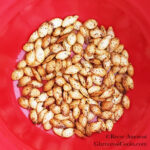 This square photo shows the completed Roasted Squash Seeds recipe by Reese Amorosi for GlamorosiCooks.com. The squash seeds are in a round formation in a red bowl. The photo is taken close so the red goes all the way to the edges of the frame. From a distance it looks like a light yellow circle in a bright red square. Up close, the seeds are oval with a pointy end and a rounded end. They are pale yellow tinged with orange and caramel brown from being roasted. They look spotty due to being seasoned with smoked paprika.
