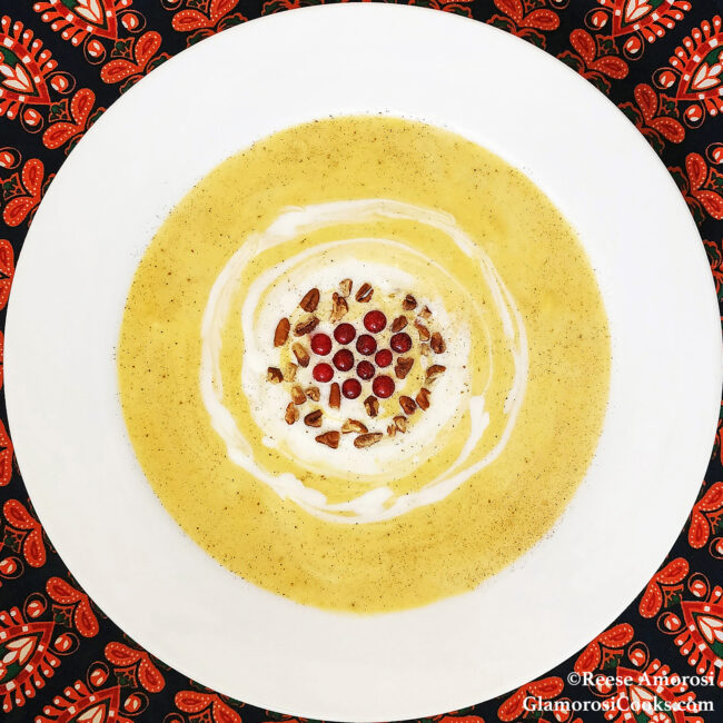 This square photo shows the completed Butternut Squash and Apple Soup recipe by Reese Amorosi of GlamorosiCooks.com. The round bowl is white and the soup is pale orange. The soup is garnished with a swirl of maple creme fraiche, then topped with pecans, red currants and a dusting of nutmeg. The bowl is in closeup, but around the edges there is a a tablecloth with a black, red, orange and white printed design.