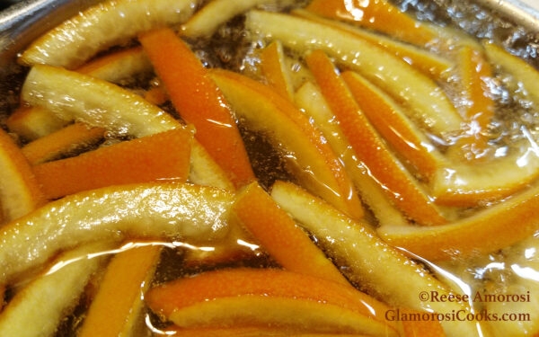 This rectangle photo shows part of the process of the Candied Orange Peel recipe by Reese Amorosi at Glamorosi.com. It is a close-up of strips of orange peel in a pot - they are simmering in simple syrup (sugar and water). The outside of each peel is deep orange, the pith is pale orange. Once they are done they will be used to garnish desserts and cheese trays.