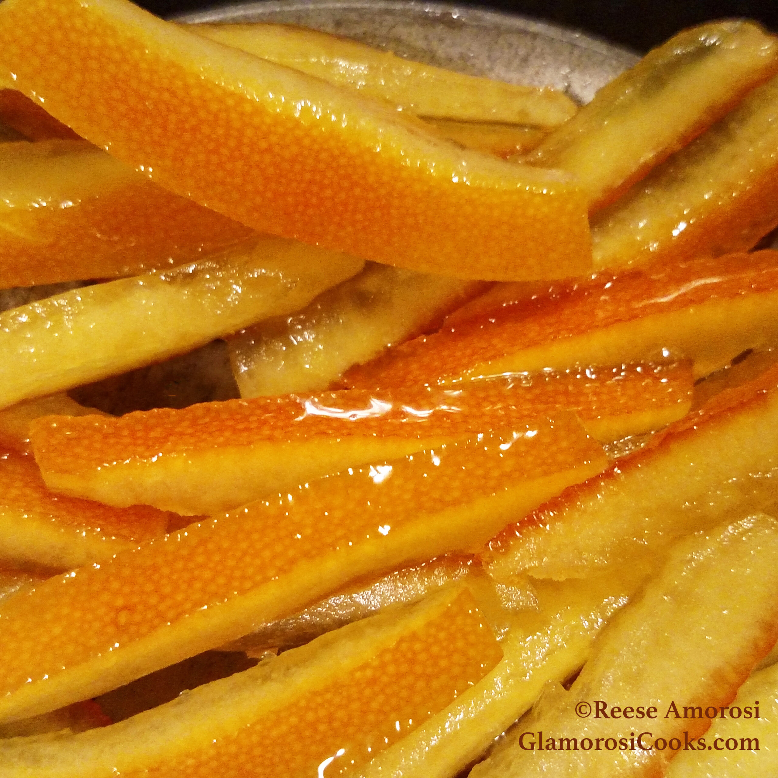 This square photo shows the completed Candied Orange Peel recipe by Reese Amorosi at Glamorosi.com. It is a close-up of strips of orange peel that have been candied by cooking them in simple syrup (sugar and water). The outside of each peel is deep orange, and the pith (the inside part of the peel) is pale orange. They will be used to garnish desserts, baked good, drinks and cheese trays.