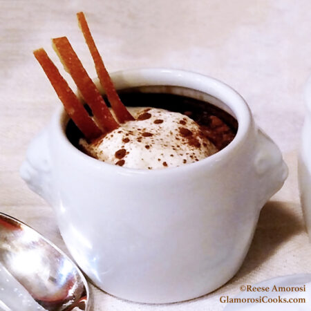 This square photo shows the completed Chocolate Chai Pots de Crème recipe by Reese Amorosi for GlamorosiCooks.com. The chocolate custard is in a white ceramic pot with lion heads for handles. The chocolate is topped with homemade whipped cream and a dusting of cinnamon. Three candied orange peels are in the custard on the left side. Part of a spoon is visible in the bottom-left corner, and the small white pot lid is barely visible in the bottom-right corner.