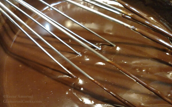 This photo shows uncooked Chocolate Chai Pots de Crème by Reese Amorosi for GlamorosiCooks.com. A whisk is in the chocolate and stretches across the entire photo on a downward diagonal from the top-left corner to the bottom-right corner.