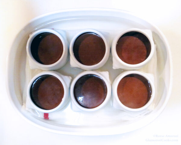 This photo shows part of the process of making Chocolate Chai Pots de Crème by Reese Amorosi for GlamorosiCooks.com. There are six pot de crème cups filled with the chocolate custard and the are sitting in a water bath inside of a white oval baking dish.