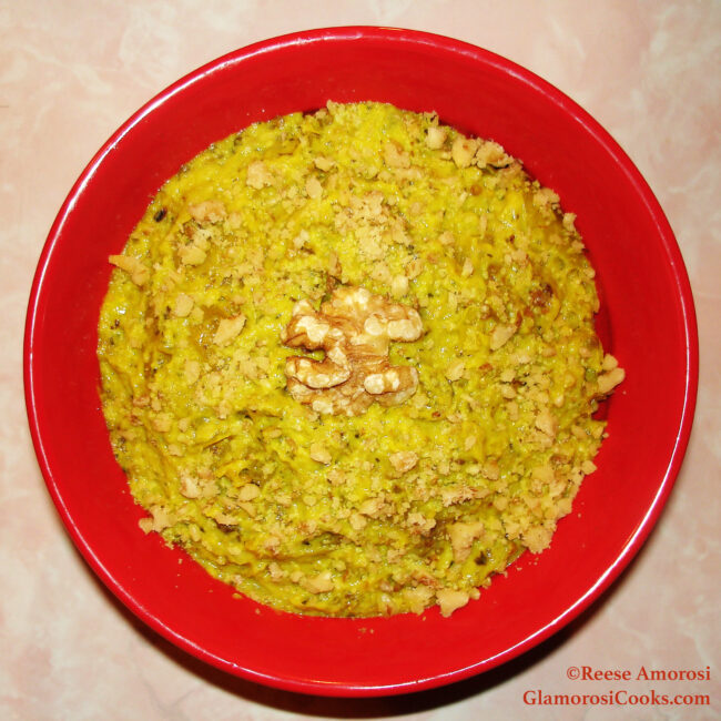 This square photo shows the completed recipe for Roasted Eggplant Dip with Yogurt, Walnuts and Mint by Reese Amorosi for GlamorosiCooks.com. The dips is yellow (due to containing turmeric) with light brown chopped walnuts, and there is a whole walnut in the middle. The dip is in a round, red bowl on a pink marbled 50s Formica dinette.