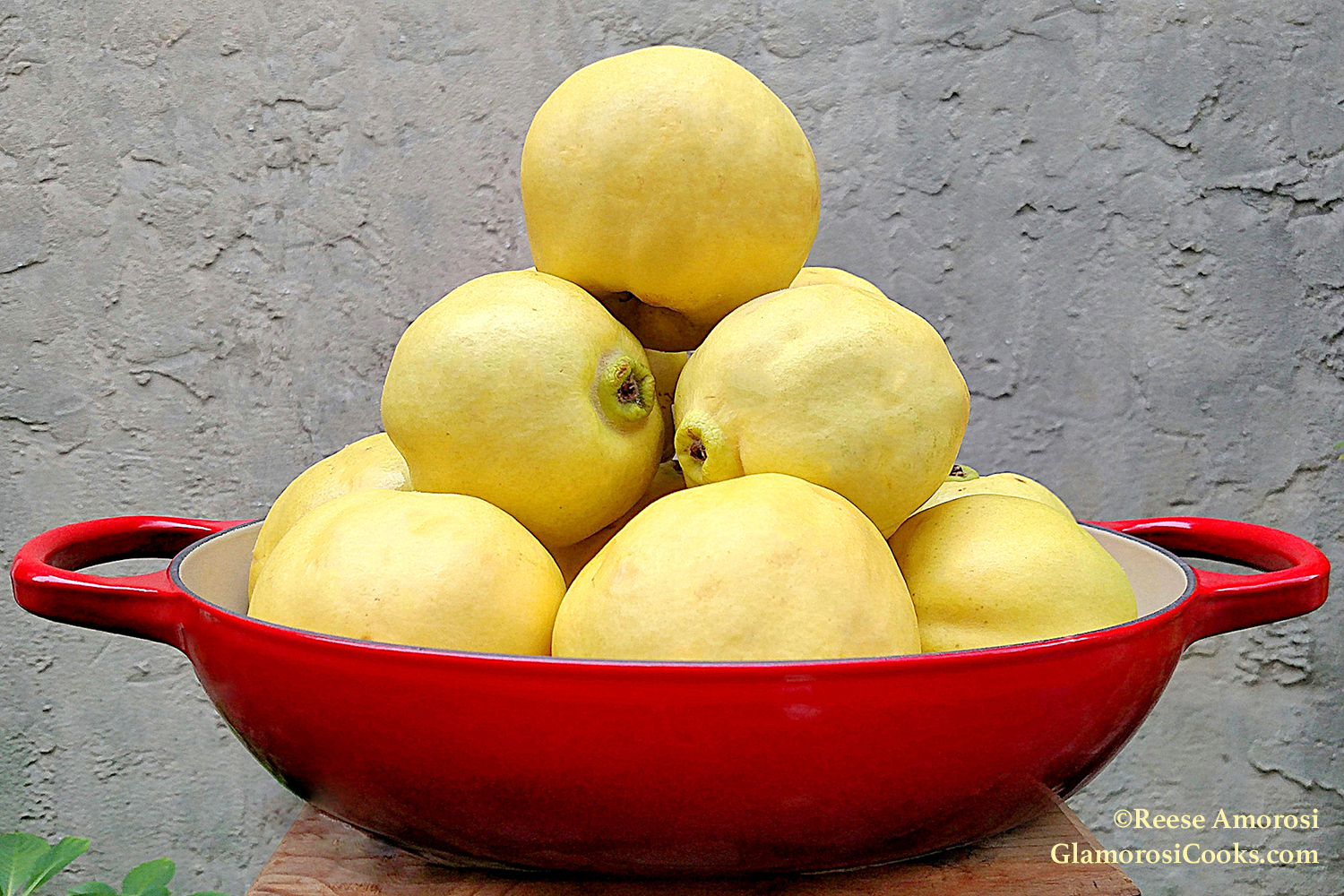 This photo is posted on the "About" page on GlamorosiCooks.com. It is a horizontal photo showing a red pan containing quince arranged in a pyramid formation. The pan is on a wood stool, and the background is a grey stucco wall. 
