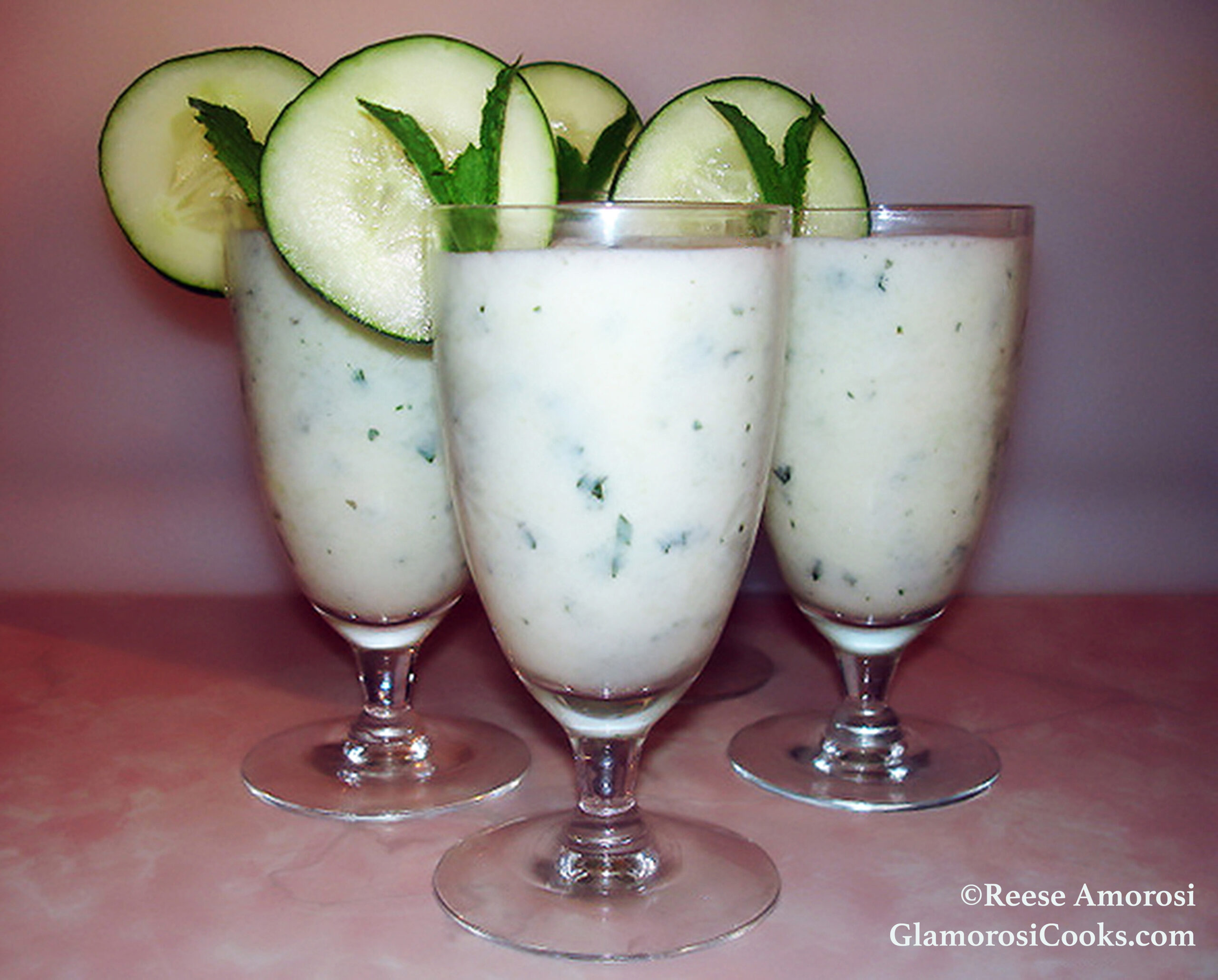 This photo shows the completed recipe for Chilled Cucumber Soup by Reese Amorosi for GlamorosiCooks.com. The soup is an off-white color with green spearmint leaves in the mix. It is shown served in cocktail glasses; all four of them are garnished with round cucumber slices and a sprig of mint.