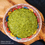 This photo shows the completed recipe for Classic Basil Pesto by Reese Amorosi for GlamorosiCooks.com. The pesto is a green sauce and in the photo you can see the texture of the combined ingredients. The pesto is in a brown ceramic bowl with a colorful floral edge. The bowl is centered on a freeform wood serving board, and that sits upon a dark blue tablecloth. The serving board cuts across the photo diagonally from top-left to bottom-right.