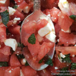 This photo shows the completed recipe for Watermelon, Feta and Tomato Salad with Mint by Reese Amorosi for GlamorosiCooks.com. The ingredients are cut small but the photo is close up. A vintage spoon containing some of the salad cuts vertically through the center of the photo.