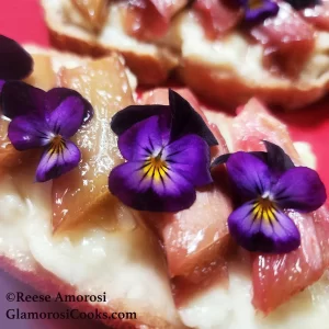 This square photo shows the completed recipe for Honey-roasted Rhubarb Toast with Brie by Reese Amorosi on the GlamorosiCooks.com website. The photo shows toast covered in brie cheese topped with honey-roasted rhubarb garnished with purple violas.