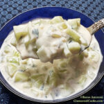 This photo shows the finished recipe for the Cucumber, Yogurt and Mint Salad Recipe by Reese Amorosi for GlamorosiCooks.com. The salad is shown in a blue bowl atop a floral light and dark blue tablecloth with a spoonful of the salad held above the bowl.