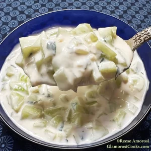 This photo shows the finished recipe for the Cucumber, Yogurt and Mint Salad Recipe by Reese Amorosi for GlamorosiCooks.com. The salad is shown in a blue bowl atop a floral light and dark blue tablecloth with a spoonful of the salad held above the bowl.