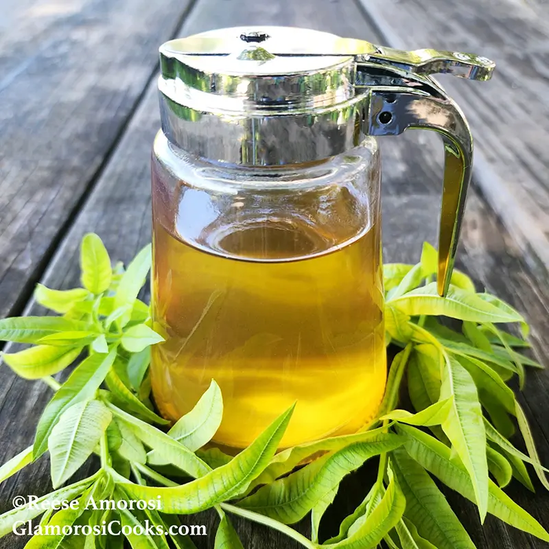 This photo shows the completed recipe for Herbal Simple Syrup, Lemon Verbena by Reese Amorosi for GlamorosiCooks.com. The clear, golden syrup is in a small glass pitcher on a wooden table. The base of the pitcher is surrounded by fresh Lemon Verbena leaves.