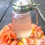 This photo shows the completed recipe for Rose Simple Syrup by Reese Amorosi for GlamorosiCooks.com. The clear pink syrup is in a small glass pitcher on a wooden table. The base of the pitcher is surrounded by a rose and rose petals.