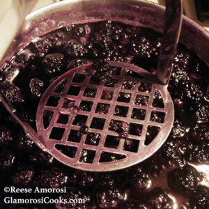 Blackberry Syrup Recipe by Reese Amorosi for GlamorosiCooks.com. In this photos the berries are in a pot and are being broken down with a potato masher. 