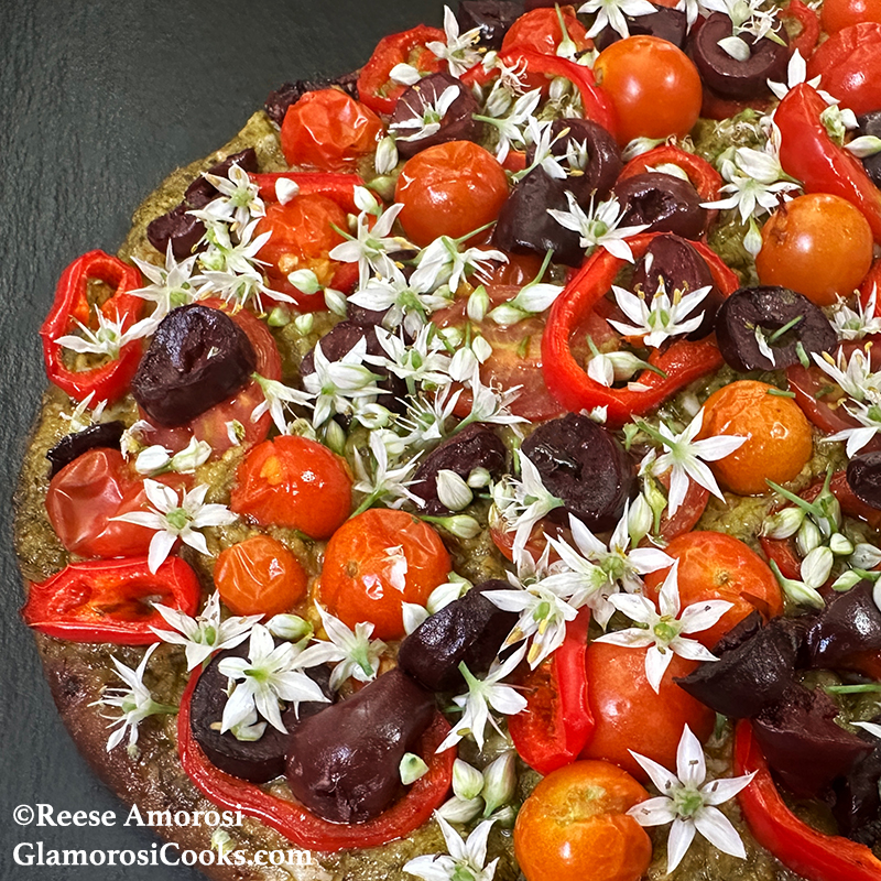 This photo shows the completed recipe for Easy Flatbread Pizza by Reese Amorosi for GlamorosiCooks.com. The flatbread is topped with pesto, cheese, tomatoes, peppers, olives and garlic chive flowers.