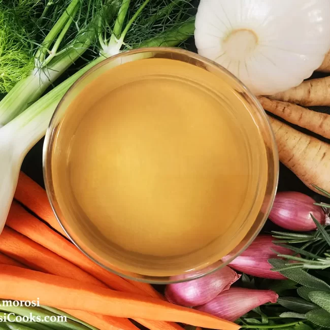 This photo shows the completed recipe for Vegetable Stock by Reese Amorosi for GlamorosiCooks.com. There is a bowl of stock surrounded by assorted vegetables and herbs.