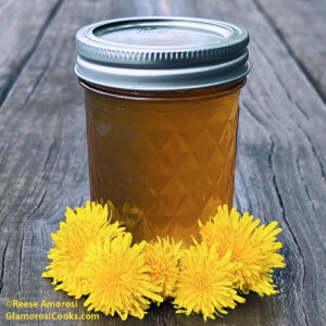 This square photo is part of the Dandelion Jelly Recipe on the Glamorosi Cooks website. The photo shows a jar of dandelion jelly on a wooden picnic table. There are five dandelion blossoms at the base of the jar. Photo ©Reese Amorosi GlamorosiCooks.com