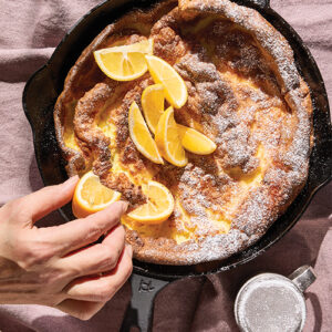 This photo featured in a review at GlamorosiCooks.com is from the cookbook MORNING BAKER (W. W. Norton & Company) by Roxana Jullapat with Ari Smolin. It shows a Dutch Baby in a cast iron pan. It is topped with powdered sugar and lemons, and a hand is shown squeezing one of the fruit wedges. Photo by Kristin Teig.